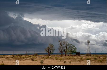 Dramatic storm clouds gather over a rural landscape with a windmill and barren trees. Stock Photo