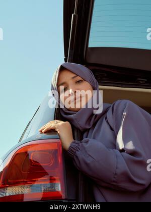 Woman in a hijab leaning on the back of a car with a blue sky in the background. Stock Photo
