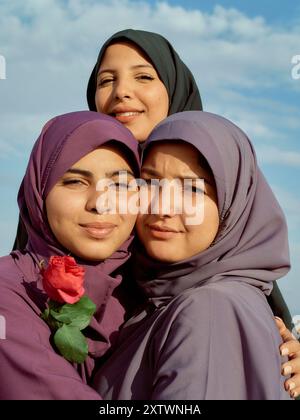 Three smiling women wearing hijabs embrace against a blue sky, one holding a red rose. Stock Photo