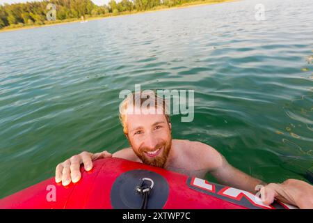 A smiling bearded man holding a red kayak in a sunlit lake. Stock Photo