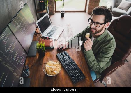 High angle view portrait of professional hacker young man eat chips computer desk loft interior office indoors Stock Photo