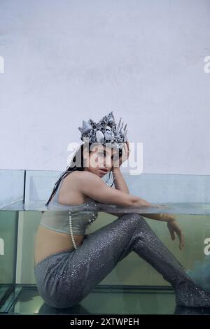 A young woman, dressed as a mermaid, sits in a glass tank, wearing a crown of shells and pearls. Stock Photo