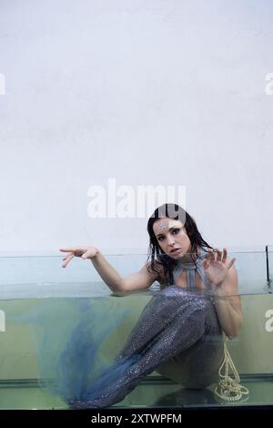 A young woman with dark hair, dressed as a mermaid, sits in a glass tank, her eyes pleading with the viewer. Stock Photo