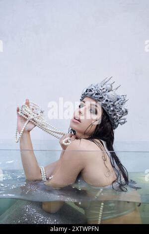 A young woman dressed as a mermaid with a seashell crown sits in a water tank, holding a pearl necklace. Stock Photo