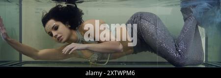 A young woman with long brunette hair swims in a large aquarium, dressed as a mermaid. Stock Photo