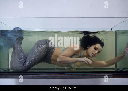 A young woman in a mermaid costume swims underwater in a large glass tank, her expression wistful. Stock Photo