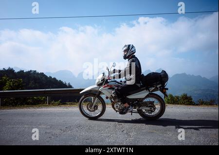 A motorcyclist in full gear rides a white adventure motorcycle on a mountain road under a clear sky. Stock Photo