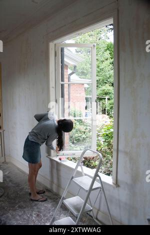 A woman in casual attire is renovating a house, scraping paint from a window frame in a room under construction, New Zealand Stock Photo