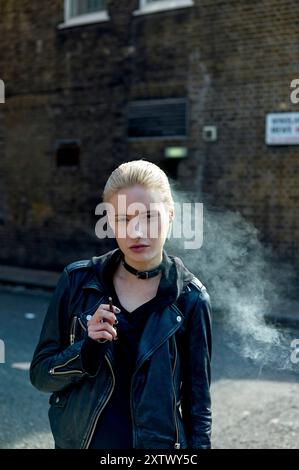 A blonde woman exhales smoke while holding a cigarette, standing in front of a brick wall with a 'No Parking' sign. Stock Photo