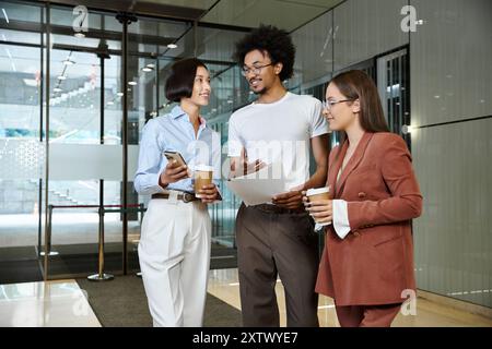 Three colleagues, stand in a glass-walled lobby, discussing work over coffee. Stock Photo