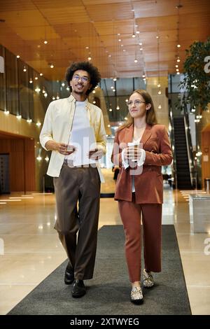 Two colleagues, a man and a woman, walk and chat in an office lobby. Stock Photo