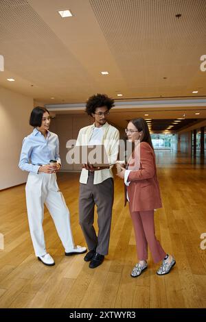 Three diverse colleagues stand in an office hallway, casually chatting and reviewing a laptop. Stock Photo