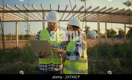 Two Caucasian experienced engineers workers woman female girl man male guy hardhat using laptop pc gadget device computer discuss talking project Stock Photo