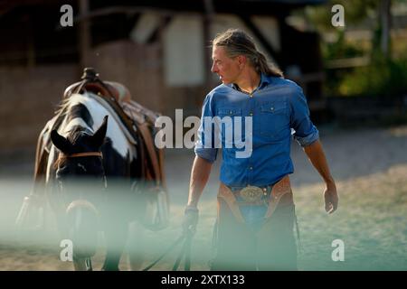 Man in a cowboy outfit standing next to a horse, holding reins in a sandy area during daytime. Stock Photo