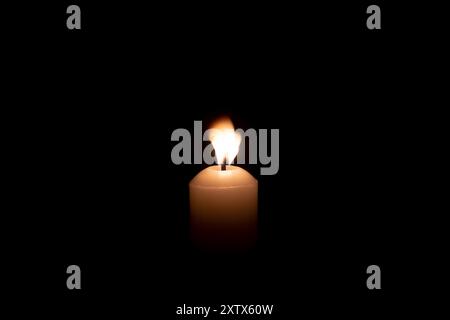 A single candle light glowing on a big white candle against blowing wind on table in church for Christmas, funeral or memorial service with copy space Stock Photo
