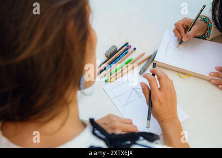 Top view, perspective of unrecognizable woman drawing on a table with notebook and colored pencils, she has a graphite pencil in her hand, there are c Stock Photo