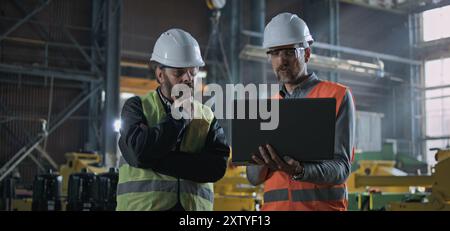 Two professional heavy industry workers, engineers wearing protective uniform and hardhats discuss work process using laptop computer while standing in manufacturing factory or warehouse. Slow motion. Stock Photo