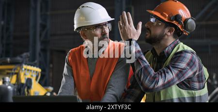 Two professional heavy industry engineers in protective uniform and helmets stand at manufacturing factory, work using laptop and talk. Mature supervisor and adult employee at plant. Slow motion. Stock Photo