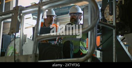 Two professional heavy industry technicians in safety uniforms and hard hats work with equipment at manufacturing factory. Mature supervisor or engineer uses laptop and talks to workers. Slow motion. Stock Photo