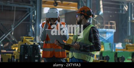 Two professional industry technicians wearing protective uniform and helmets stand at manufacturing factory. Mature engineer uses VR headset, adult employee work in laptop computer. Slow motion. Stock Photo