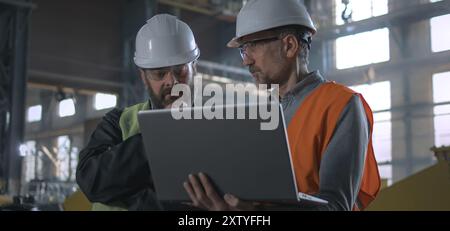 Two professional heavy industry workers, engineers wearing protective uniform and hardhats discuss work process using laptop computer while standing in manufacturing factory or warehouse. Slow motion. Stock Photo