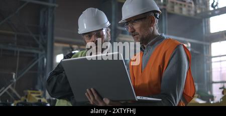 Two professional heavy industry workers, engineers wearing protective uniform and hardhats discuss work process using laptop computer while standing in manufacturing factory or warehouse. Slow motion. Stock Photo