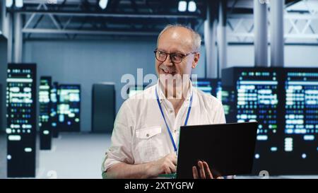 Elderly engineer expertly managing data while navigating in industrial server farm. Senior technician ensuring perfect cybersecurity protection, optimizing systems Stock Photo