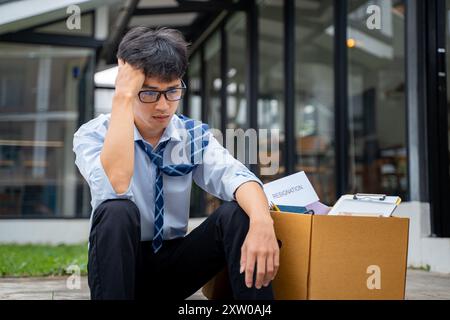 Sad young businessman sitting at a curve side road with a box full of his office stuff beside him Stock Photo