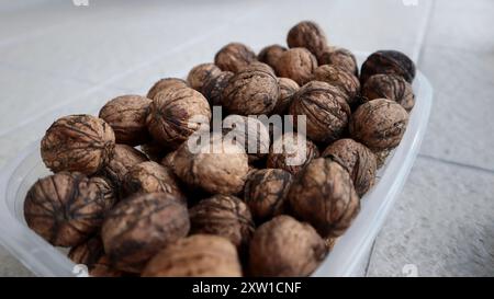 A close-up of a plastic container filled with whole walnuts. The walnuts have dark lines and strings (known as peduncles or fibers) on their shells, w Stock Photo