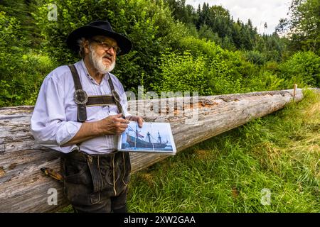 Raftsman Martin Spreng stands beside a felled Dutch fir ("Holländertanne") and shows a photograph of a historic sailing ship. Black Forest raftsmen felled particularly long, straight firs for Dutch shipbuilding – knot-free over long stretches and 30 to 40 metres long. Altensteig, Baden-Württemberg, Germany. Rafting shipped such timber here on the Nagold river until 1911. It has been recognised as UNESCO intangible cultural heritage since 2022. Stock Photo