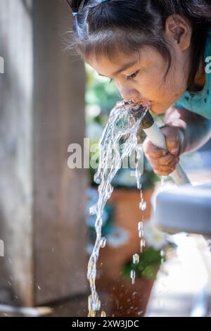 A young girl is drinking water from a hose. The water is coming out of the hose and the girl is trying to catch it in her mouth. The scene is playful Stock Photo