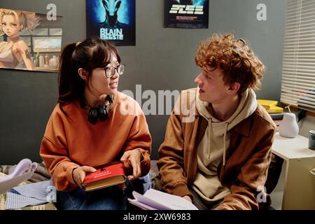 Two students sitting in cozy bedroom setting, discussing study material and holding books. Anime posters on walls and comfortable atmosphere indicating relaxed study environment Stock Photo