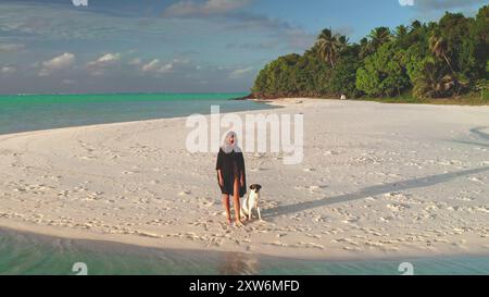 Young woman is enjoying a relaxing walk with her dog on a pristine white sand beach, with lush tropical vegetation and turquoise waters in the background Stock Photo