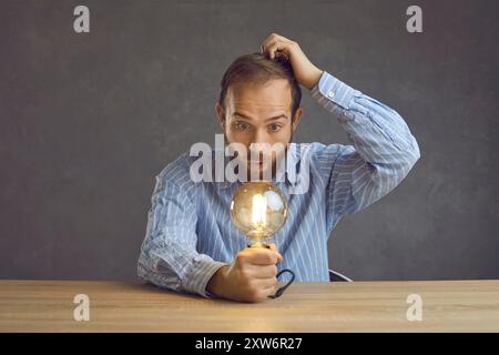 Funny man looking at shining light bulb in hand and scratching head with puzzled face expression Stock Photo