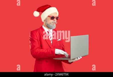 Stylish and cool man in Christmas Santa hat is using laptop with serious expression on his face. Stock Photo