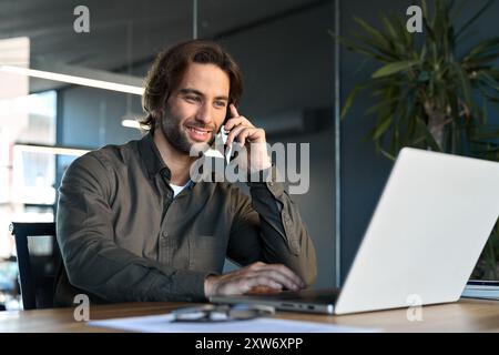 Happy business man talking on mobile phone working on laptop in office. Stock Photo