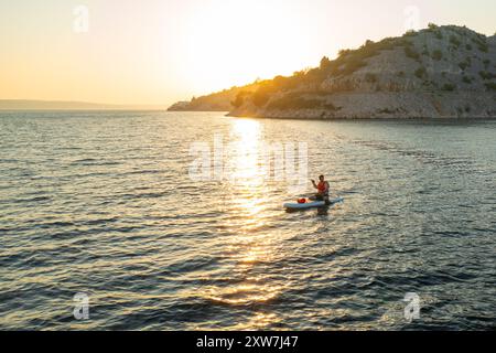 Man in an orange vest sits on his paddleboard, moving across the serene sea at sunset Stock Photo