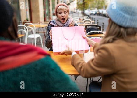 Young woman showing excitement receiving a pink shopping bag gift from friends at outdoor cafe. Autumn scene with surprised expression, warm clothing, Stock Photo
