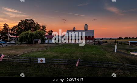 Ronks, Pennsylvania, USA, July 19 2024 - Large Red Barn With Silver Roof And Twin Cupolas At Sunset, Surrounded By Open Fields, A Weeping Willow, Fencing, And Other Farm Structures Under A Pink And Orange Sky. Stock Photo