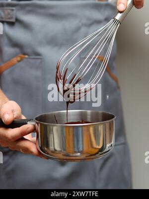 Chef making chocolate cake, closeup of hands holding a pot. Melted chocolate on a whisk. Stock Photo