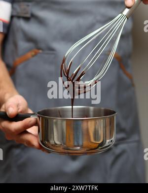 Man in grey apron with whisk and saucepan at kitchen, closeup photo of male hands. Man making chocolate. Stock Photo