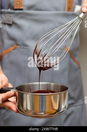 Man in grey apron with whisk and saucepan at kitchen, closeup photo of male hands. Man making chocolate. Stock Photo