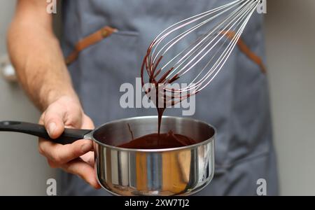 Man in grey apron with whisk and saucepan at kitchen, closeup photo of male hands. Man making chocolate. Stock Photo