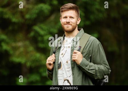 Portrait of a Caucasian man smiling while standing in forest wearing a backpack and holding its straps looking directly at camera capturing nature's essence Stock Photo
