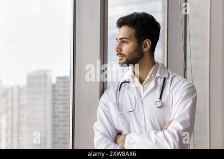 Doctor in coat standing near window, gazing thoughtfully into distance Stock Photo
