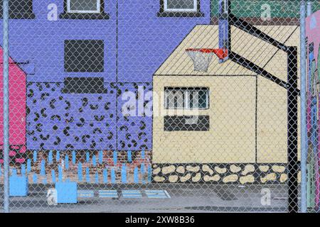 Colorful urban street art mural on building walls with a basketball hoop and playground seen through a wire chain link fence. Calgary Alberta Canada. Stock Photo