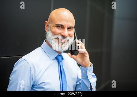 Bald bearded businessman standing next to a black wall while talking on the phone Stock Photo