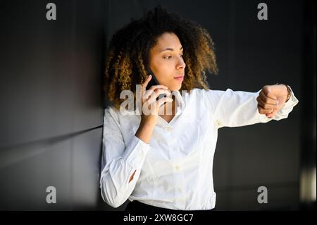 Impatient businesswoman checking watch and talking on phone in urban setting, juggling technology and communication. African american leader on the go Stock Photo