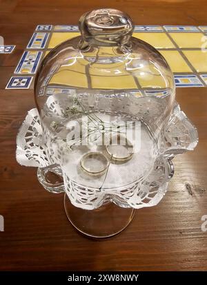 Detail of a glass dose with golden wedding rings, placed on a rustic wooden table with ceramic tiles decoration. Modra, Slovakia. Stock Photo