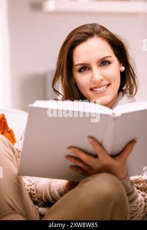 Young woman enjoying a peaceful moment at home, curled up on the sofa with a captivating book, fully immersed in reading and beaming with contentment Stock Photo